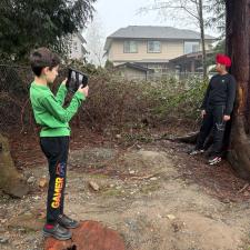 One student taking a photo of another student who is posing and leaning against a tree